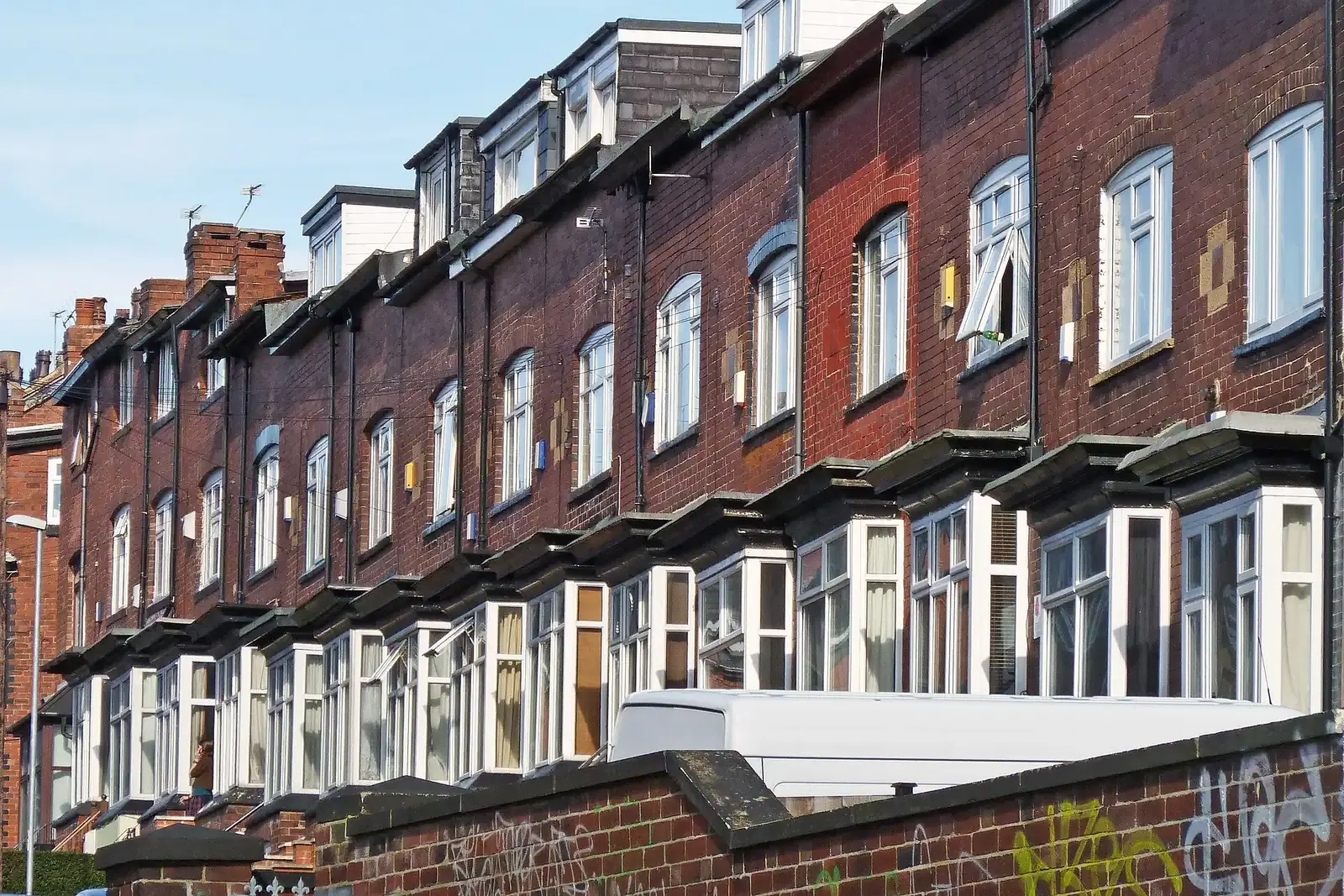 Long terrace of dark red brick houses with flat-roofed bay windows and roof dormers, climbing a hill, with a white van parked in front
