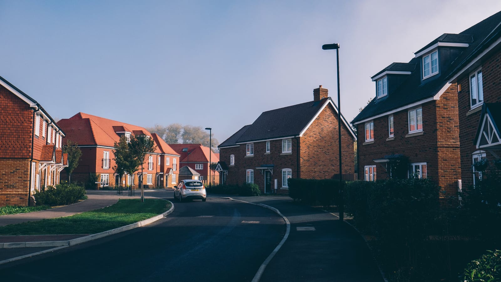 Morning view along a quiet English suburban street of red-brick houses, with front driveways opening onto the road and pavement