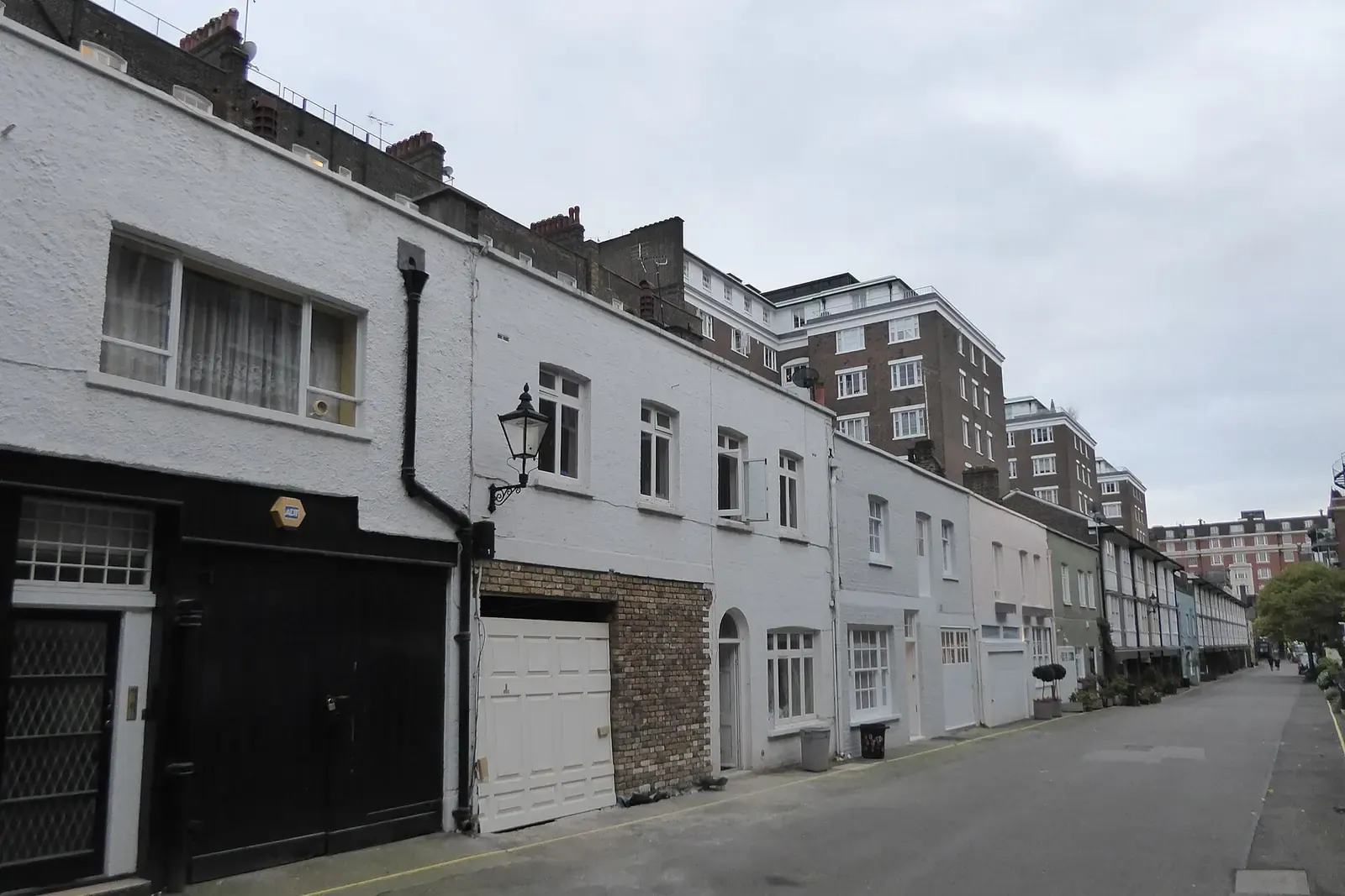 London mews of white-painted converted coach houses with black and white garage doors and a gas-style lamp, mansion flats beyond