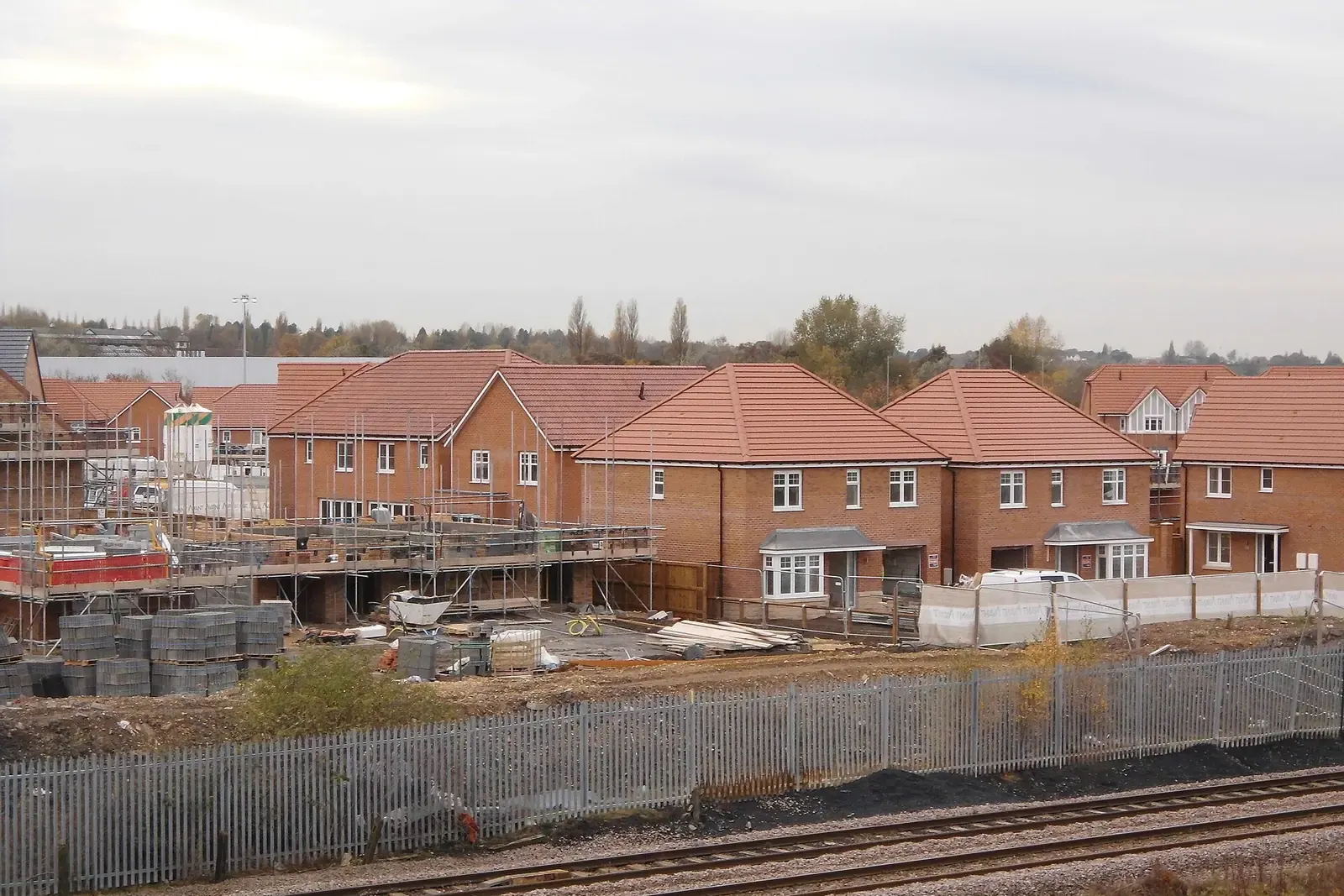 Housing estate of new red-brick houses with scaffolding and stacked blocks, behind a railway fence