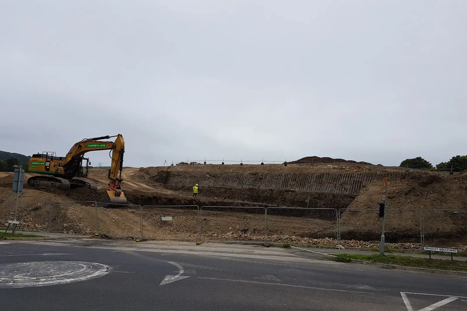 Yellow excavator working on a cut-and-fill housing site beside a roundabout, a worker in hi-vis at the edge